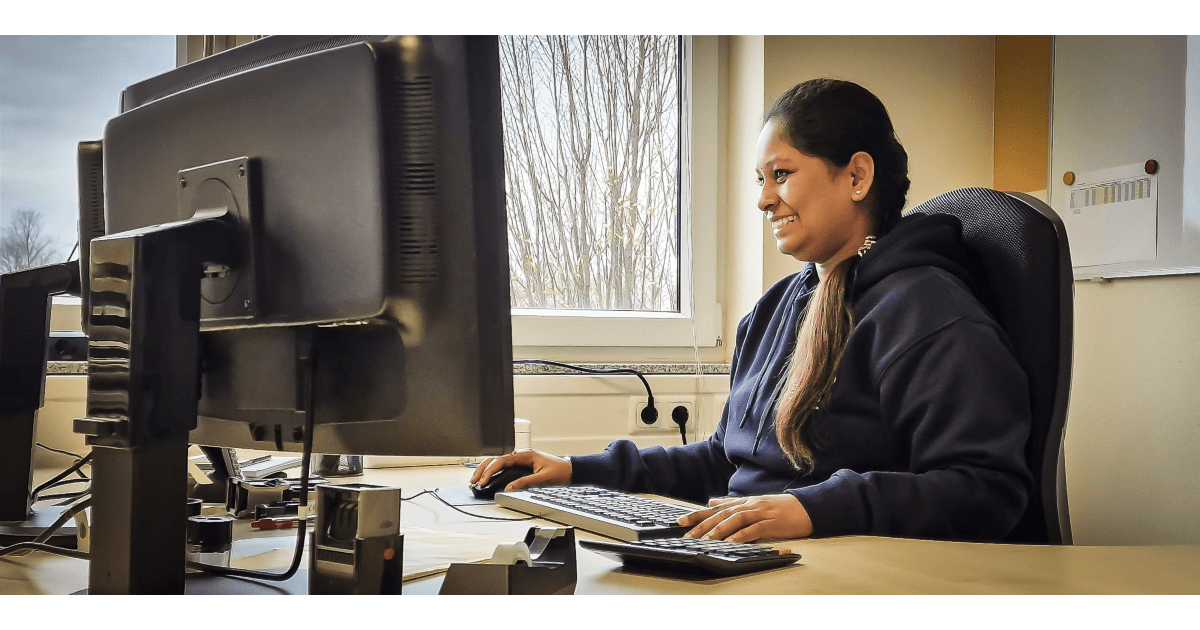 AutoGyro A Finanzbuchhalter is sitting at a desk in an office, smiling while working on a computer with dual monitors. The room has a window showing bare trees outside, and the desk is neatly arranged with office supplies like a keyboard, mouse, and organizer.